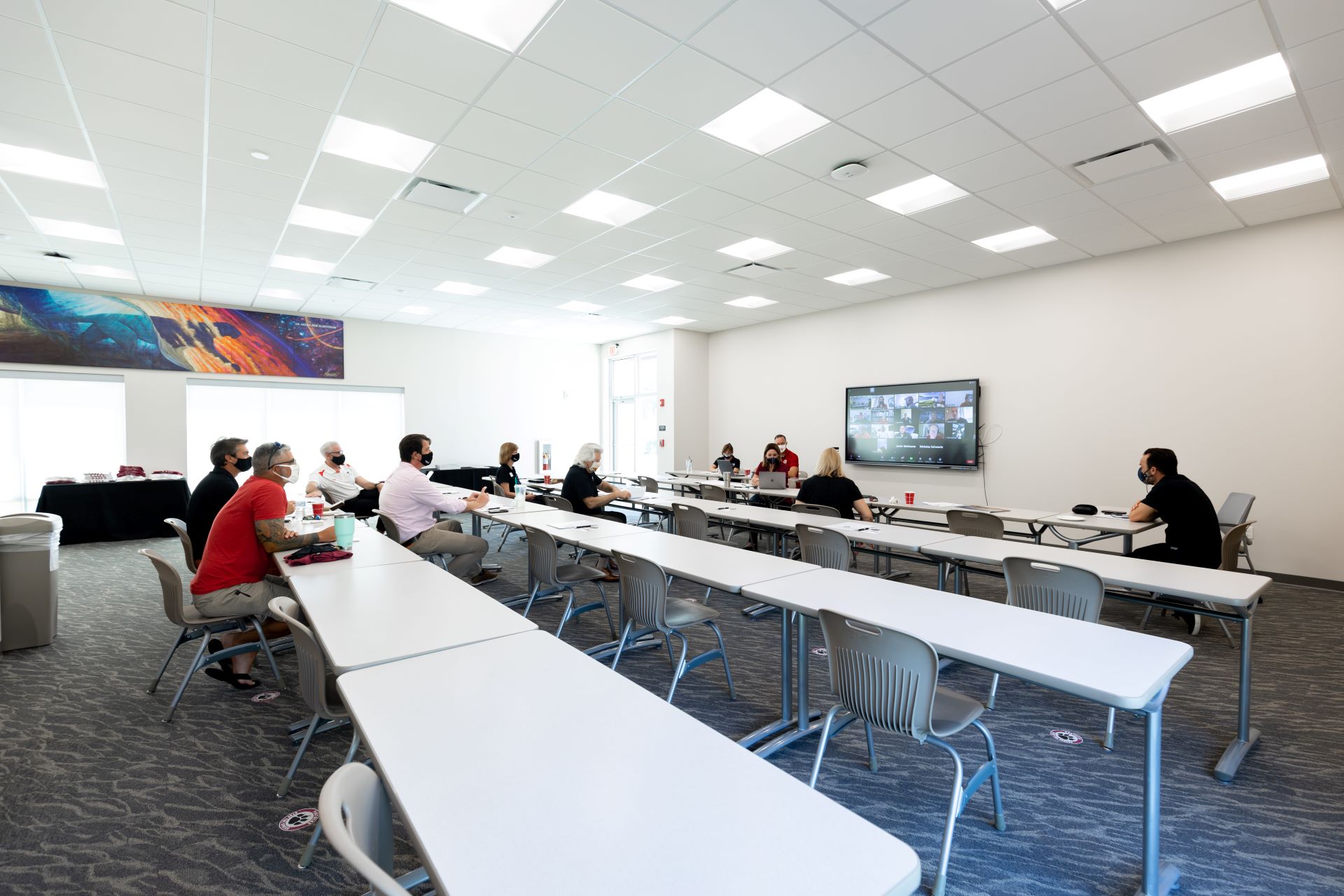 In the image, a classroom at the Folliard Alumni Center shows people wearing masks, attending a hybrid meeting with participants both in-person and via video conferencing on a large screen