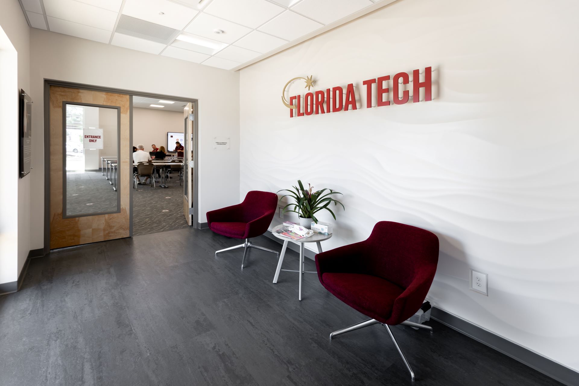 Two chairs in the Folliard Alumni Center lobby face a room where a meeting is in progress, with the 'Florida Tech' sign on the wall