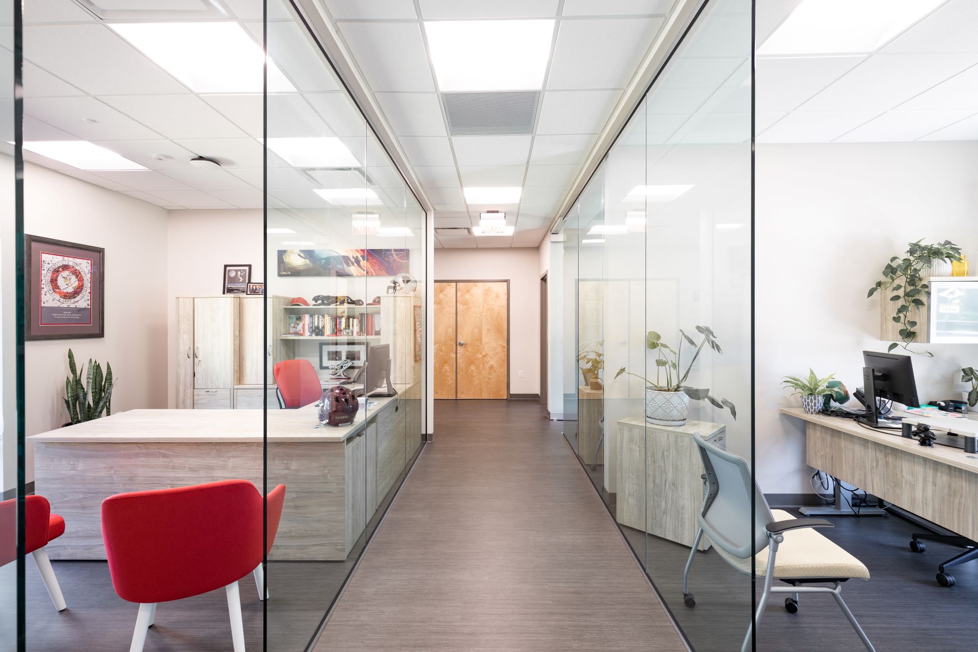 Hallway lined with glass-walled offices featuring modern wooden desks and red chairs, leading to a wooden door at the end