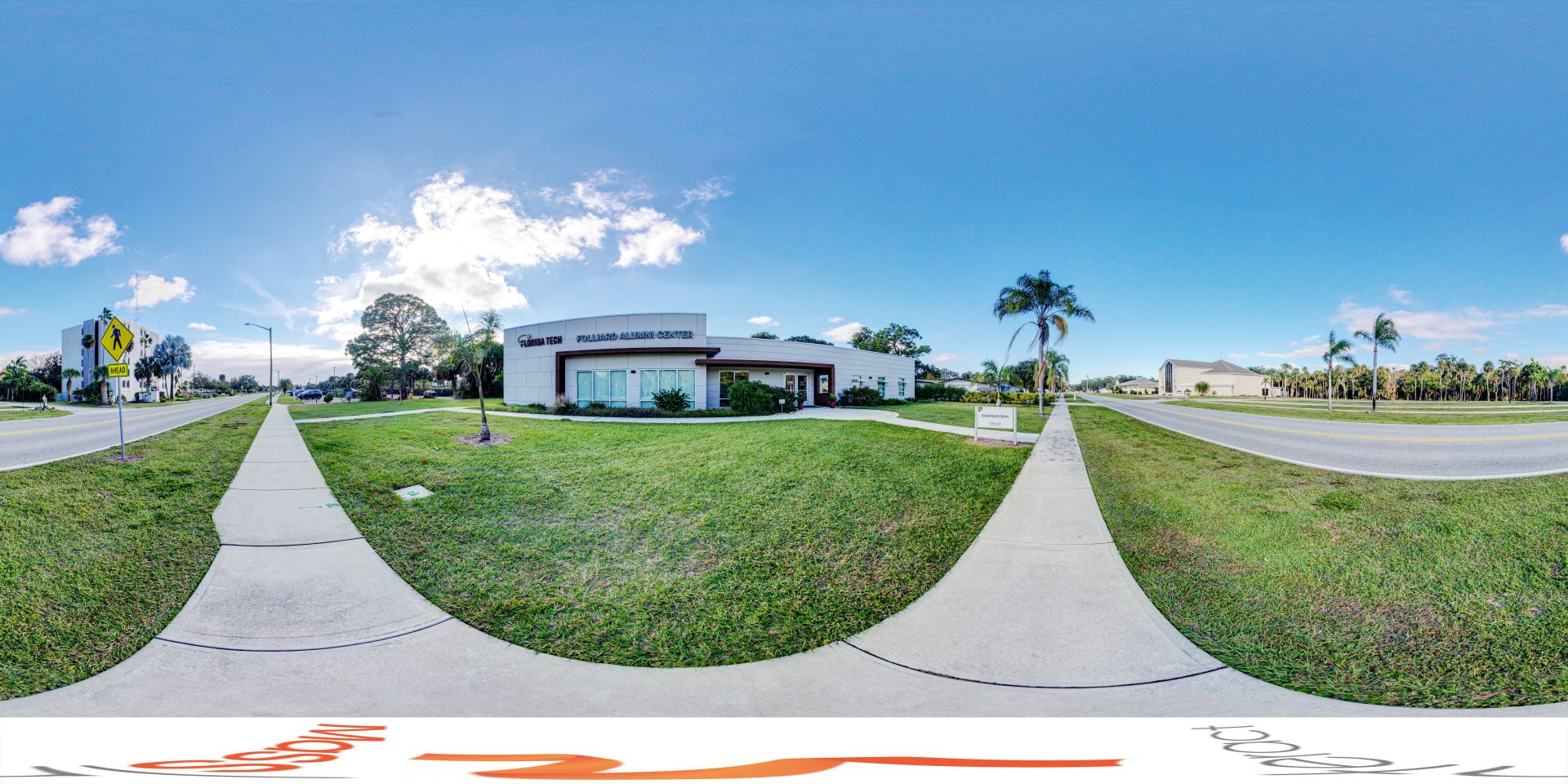Panoramic view of the Folliard Alumni Center at Florida Tech, featuring a clear sky, well-maintained lawn, and surrounding palm trees