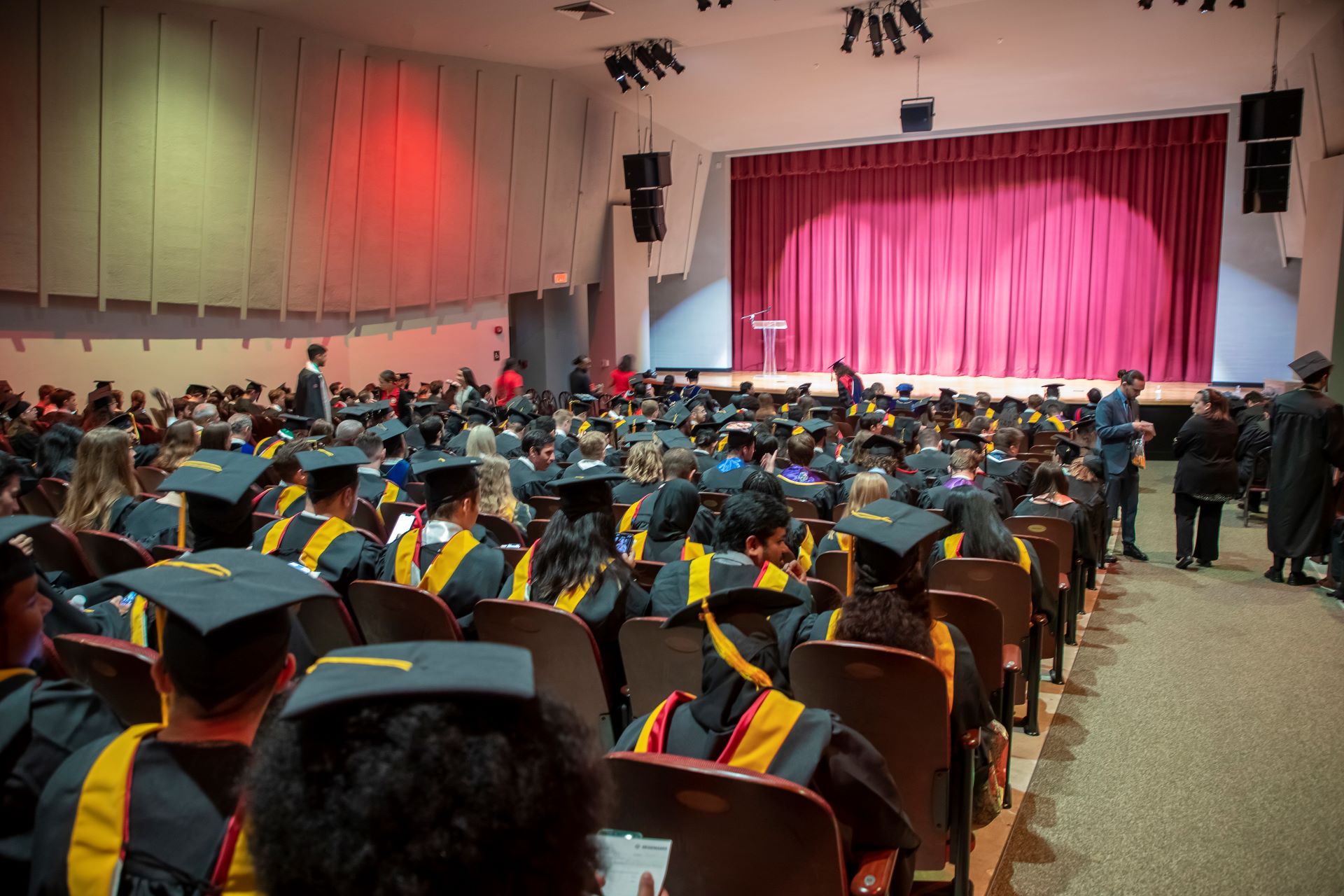 Students in graduation gowns and caps gather inside the Gleason Performing Arts Center at Florida Tech, preparing for the commencement ceremony, with the stage and red curtain in the background