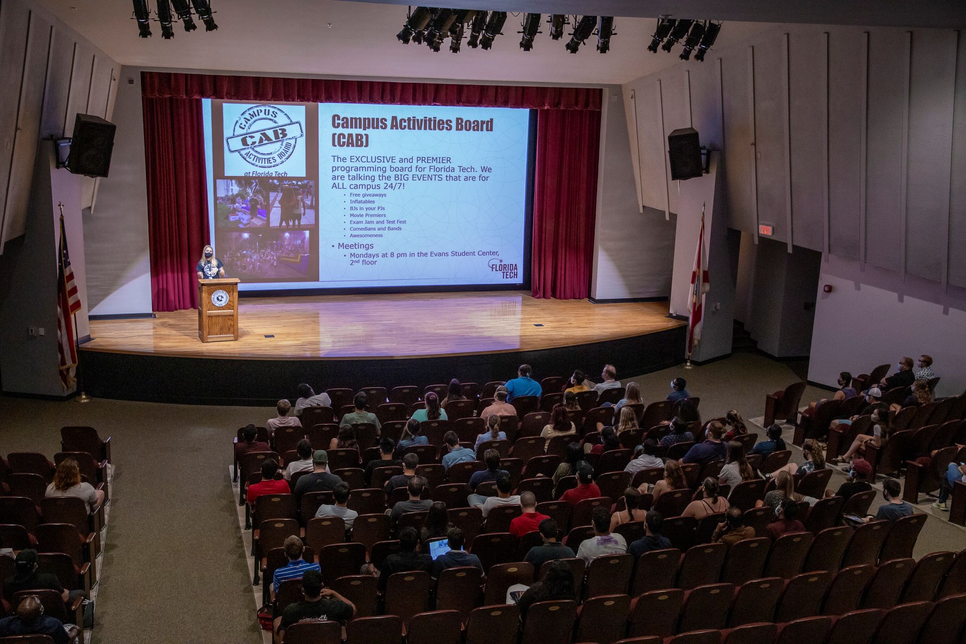 A speaker presents on stage at the Gleason Performing Arts Center, Florida Tech, to an audience, highlighting Campus Activities Board (CAB) events on a large screen