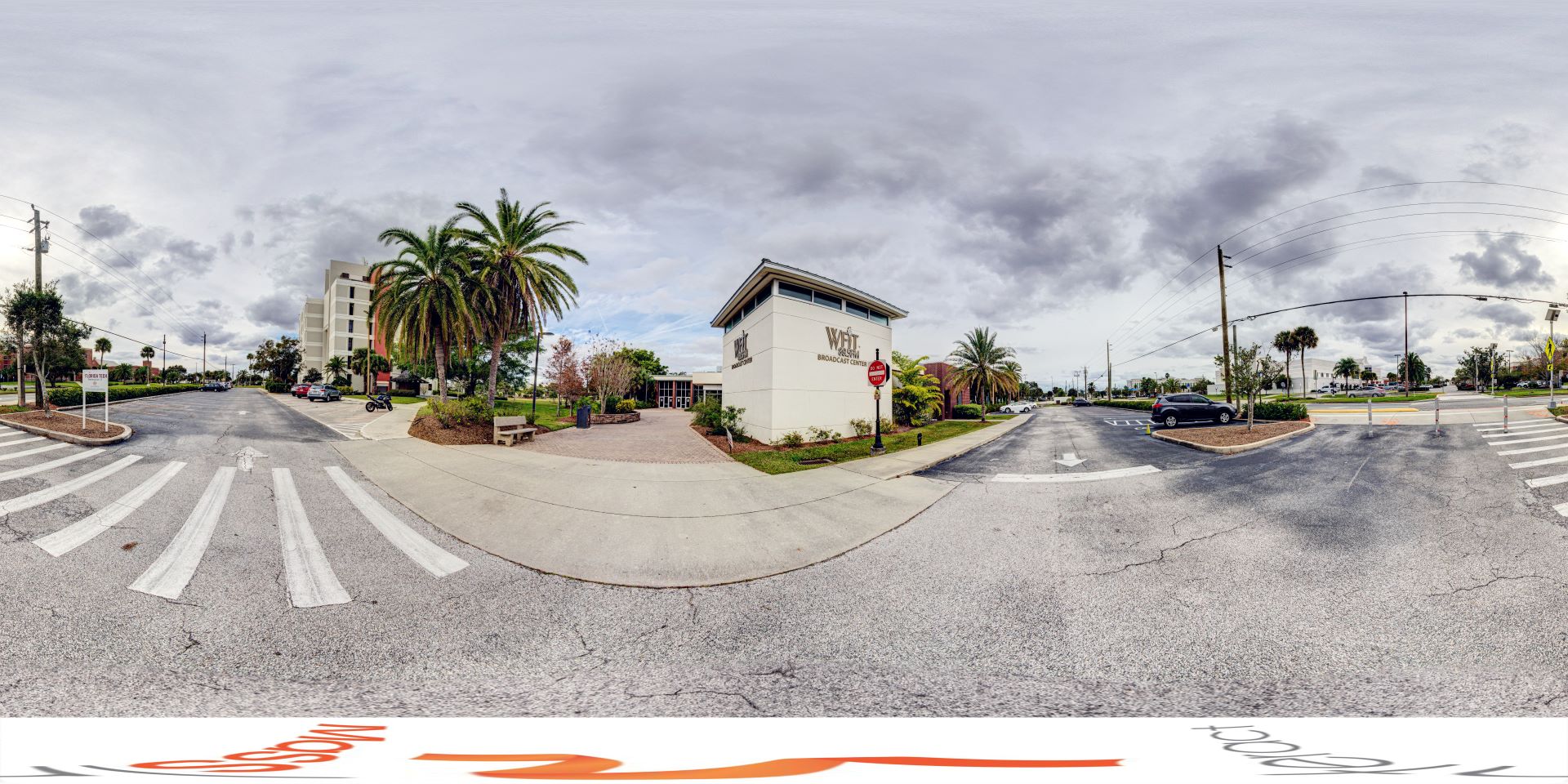 Panoramic view of the WFIT Broadcast Center located at Florida Tech, featuring the surrounding area with crosswalks, palm trees, and adjacent buildings under a cloudy sky