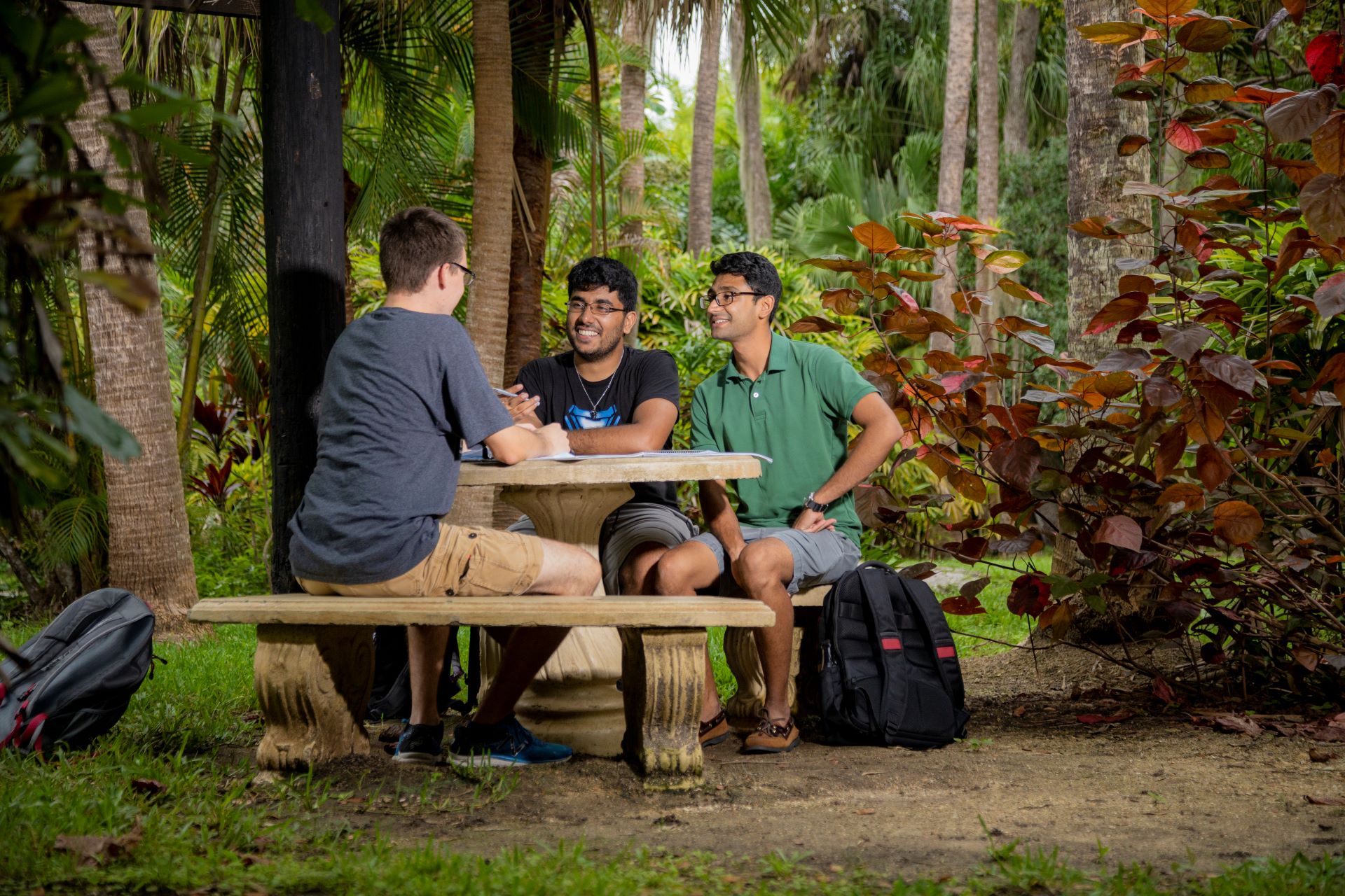 Three students sit around a stone table, engaging in conversation amidst the lush greenery of the Joy and Gordon Patterson Botanical Garden at Florida Tech
