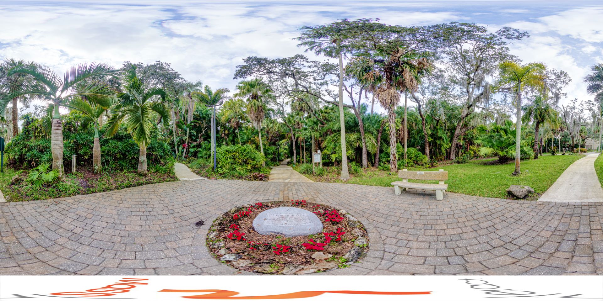 A panoramic view of the Botanical Garden at Florida Tech, featuring lush tropical vegetation, a winding path, and a commemorative plaque surrounded by flowers in a peaceful setting