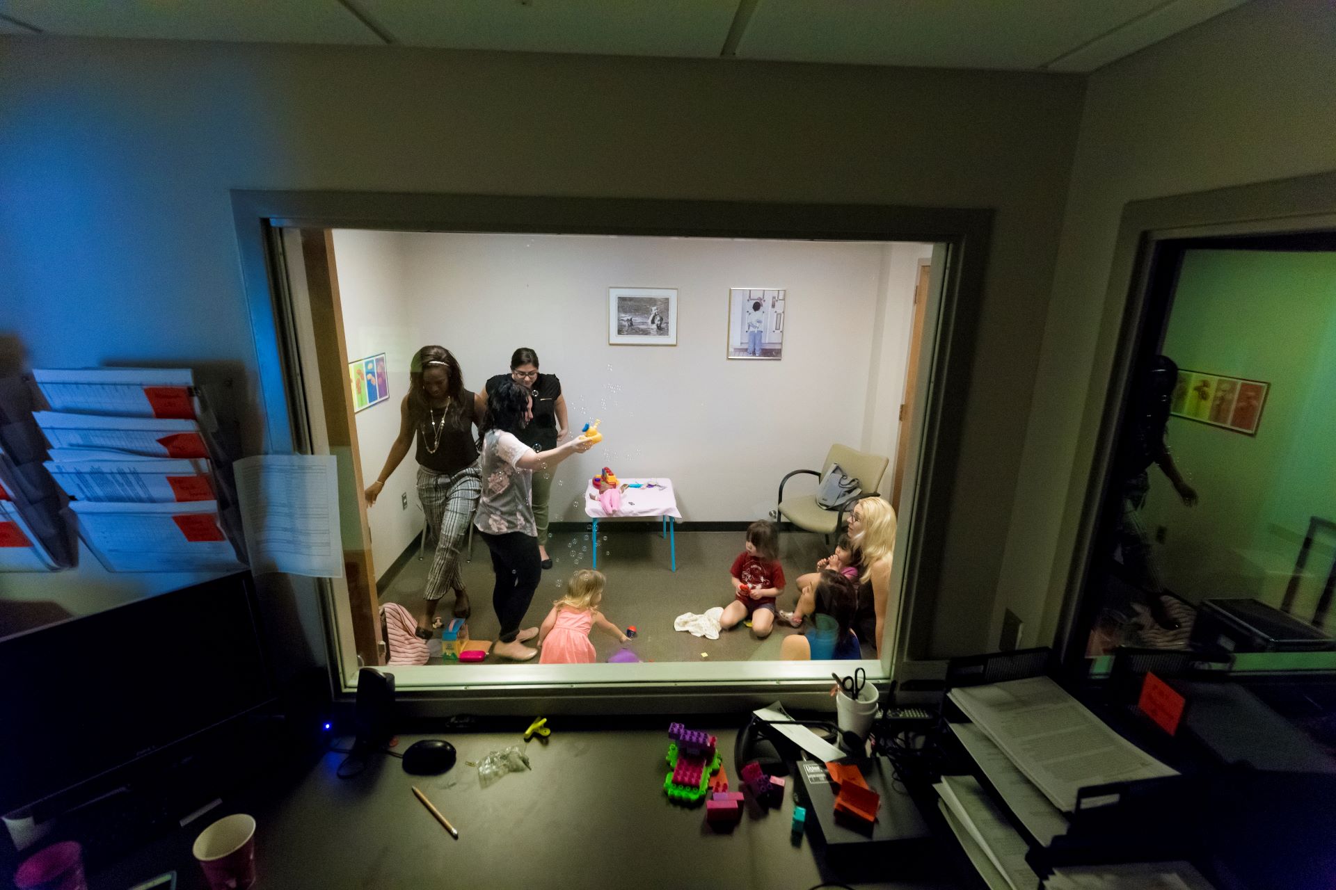 Several adults and children playing with toys in a small room observed from a larger room through a window
