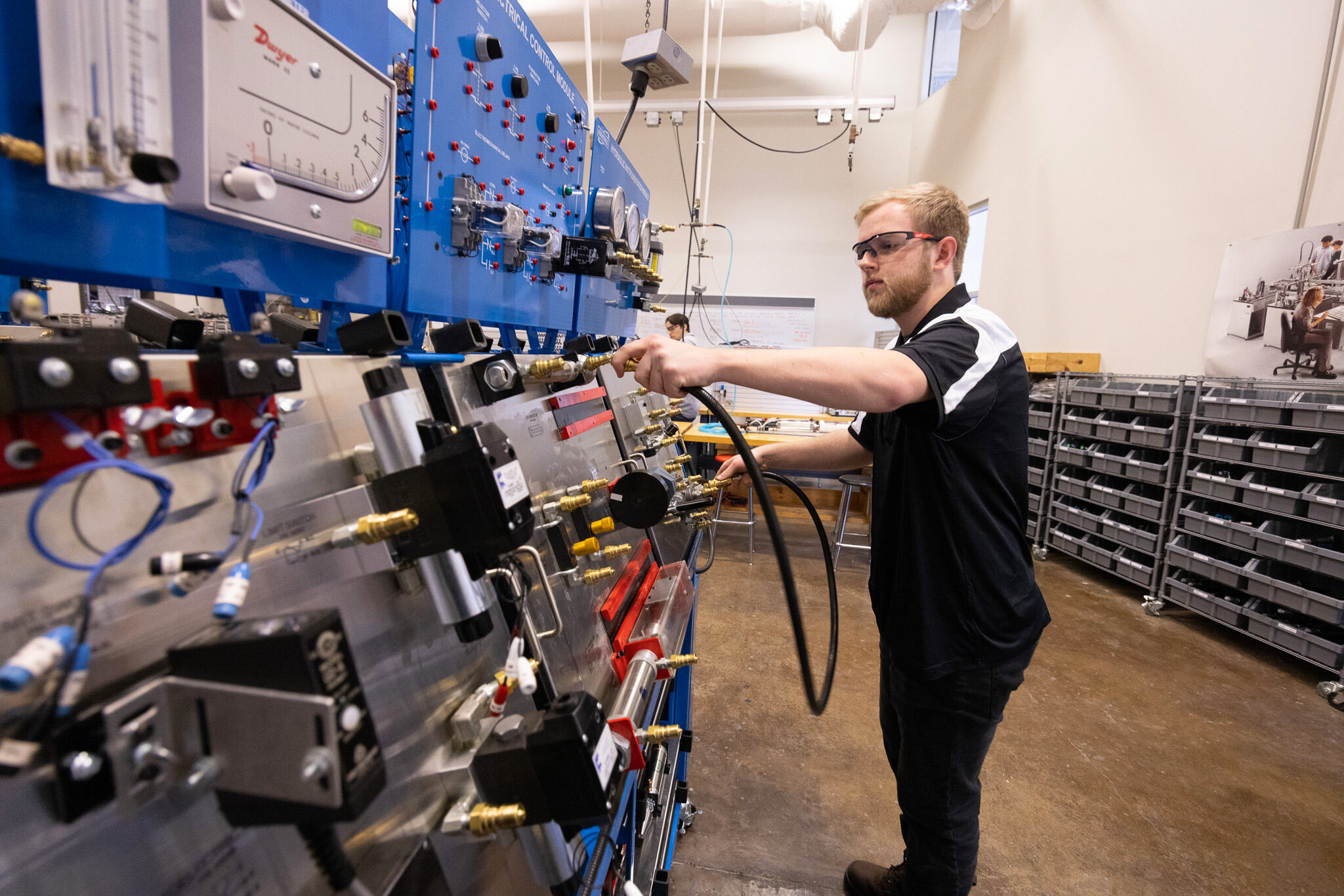 A student wearing safety glasses connects a hose to a hydraulic training panel, adjusting fittings and controls among gauges, valves, and labeled components in a lab setting.