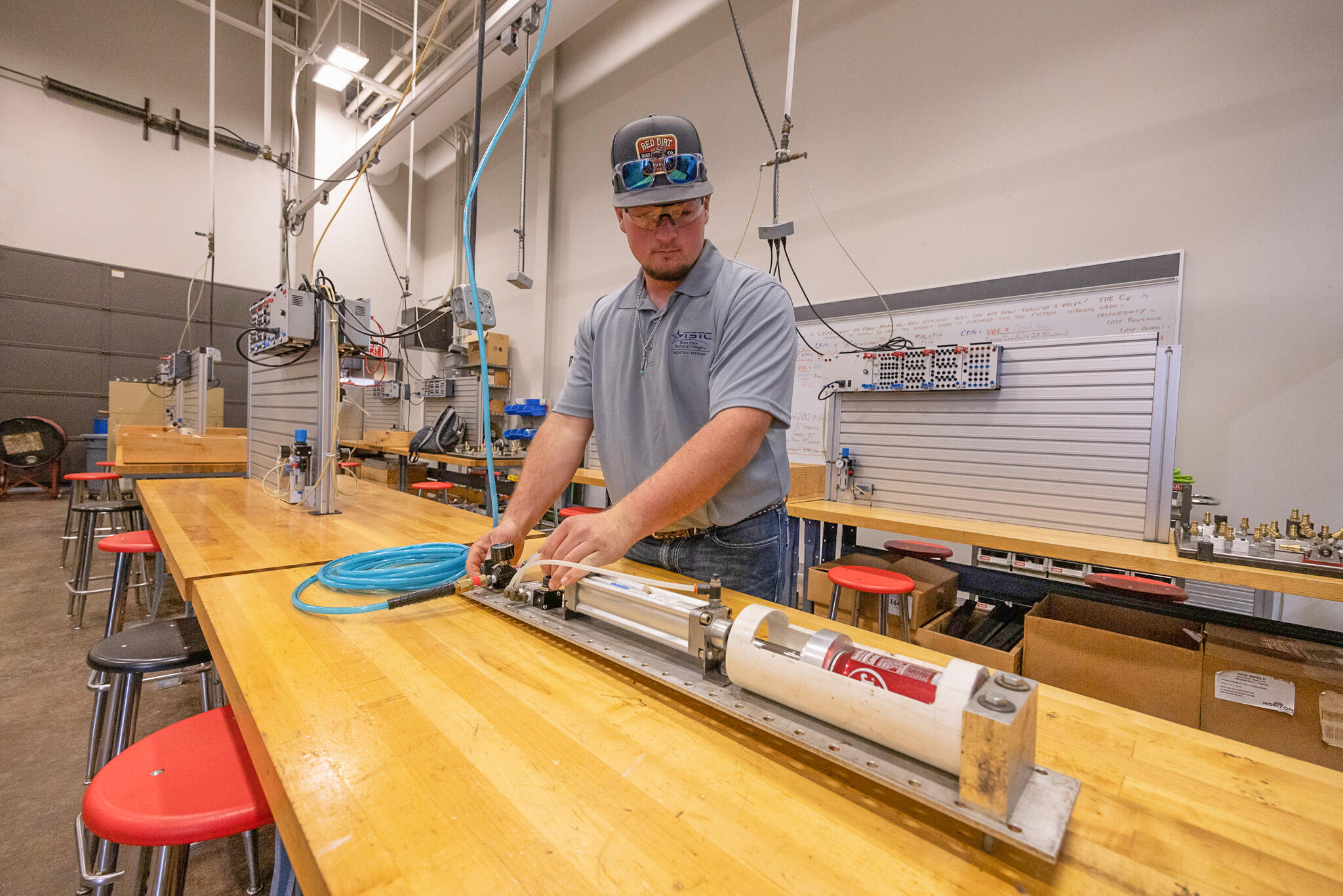 A student wearing safety glasses adjusts a pneumatic cylinder setup on a workbench, connecting hoses and fine-tuning a regulator in a hands-on lab environment.