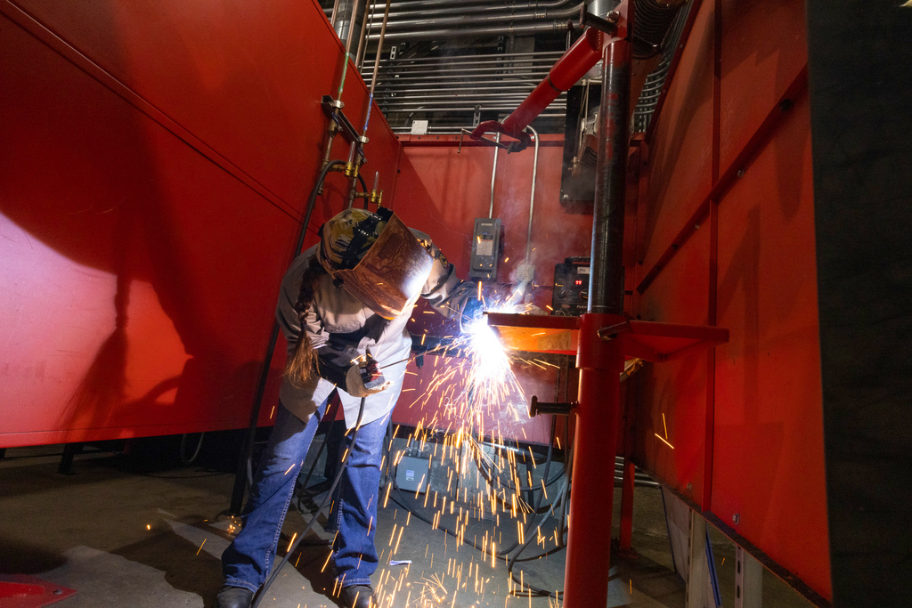 A student wearing a welding helmet and protective gear bends over a metal workpiece inside a red welding booth, creating bright sparks as they weld, with ventilation pipes and equipment visible overhead.
