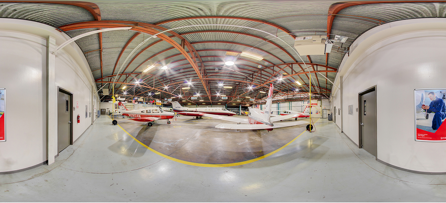 Panoramic interior of an aviation training hangar with several small airplanes parked on a polished concrete floor, surrounded by high ceilings, bright overhead lights, and doors and instructional posters along the walls.