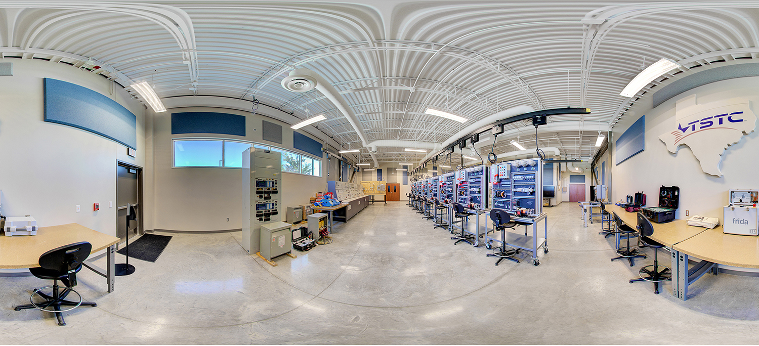 Panoramic view of a TSTC training lab with rows of electronic workstations and control panels, stools and desks arranged across a spacious room, high white ceilings with exposed piping, and instructional equipment set up for hands-on learning.