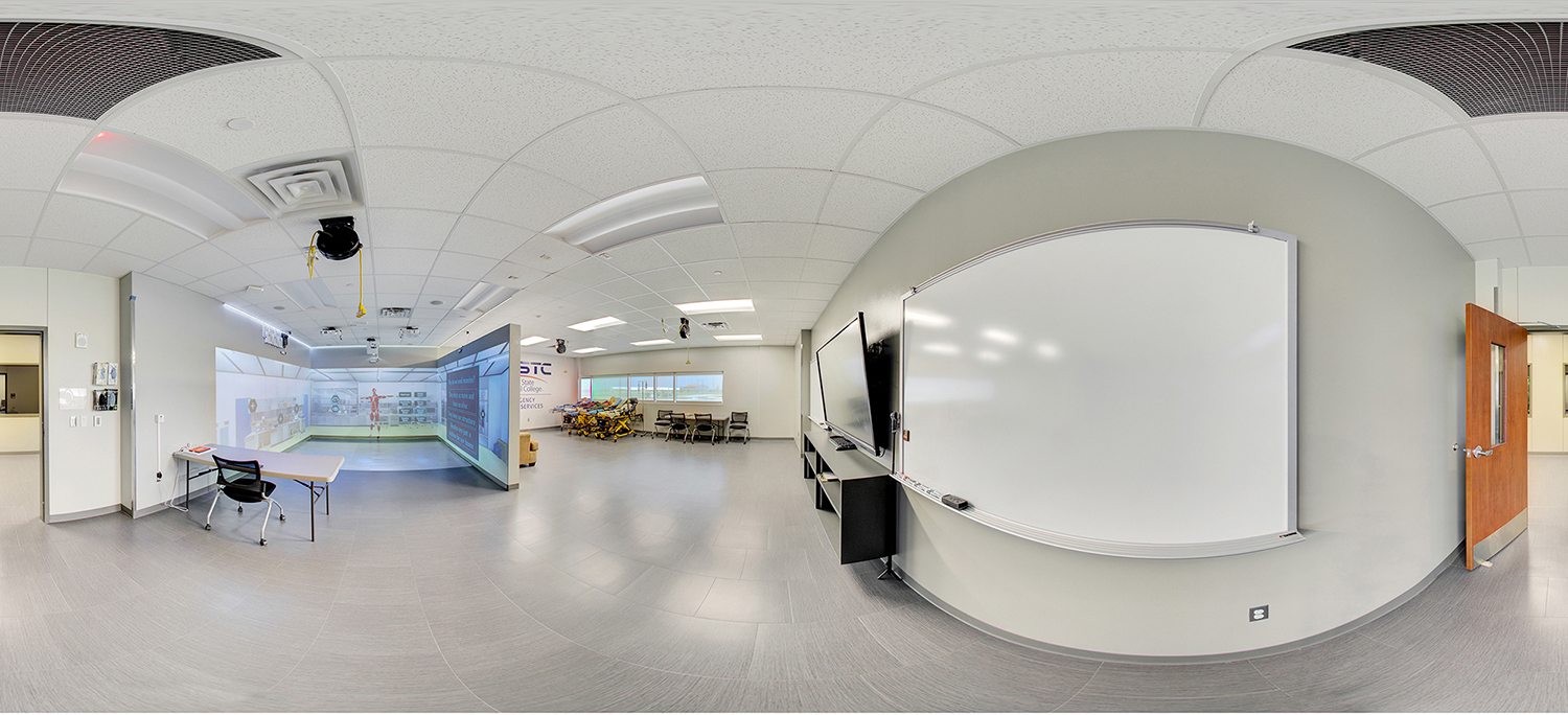 Panoramic view of a modern classroom with a large whiteboard and display screen on one side, an immersive projection training area on the other, and tables and chairs arranged for instruction under a drop ceiling with mounted equipment.