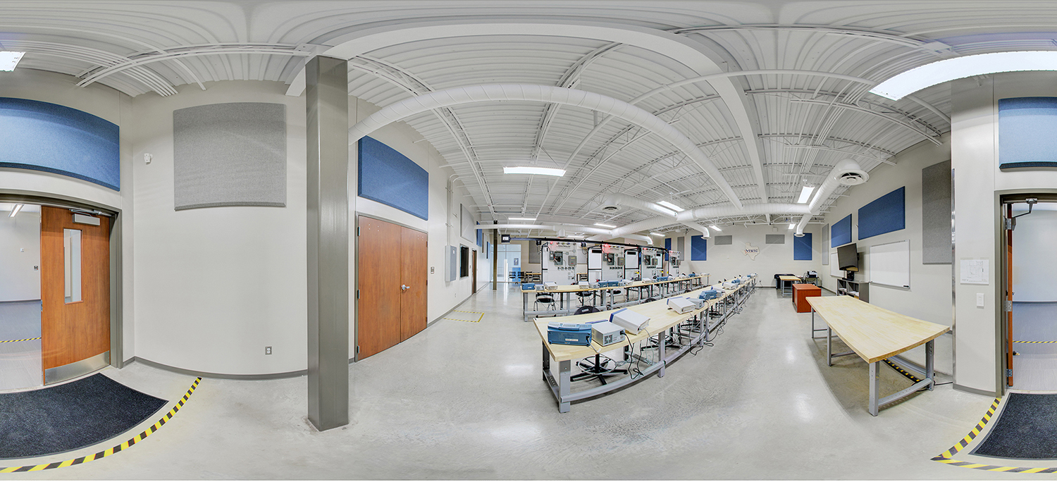 Panoramic view of a technical training classroom with long workbenches lined with equipment, stools for students, and instructional stations at the front, set beneath a high white ceiling with exposed ducts and acoustic panels.