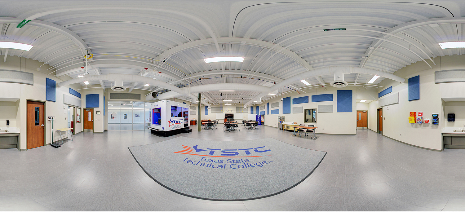 Panoramic view of a TSTC Emergency Medical Services Skills Lab training space featuring an open classroom layout with tables and chairs, instructional areas, and a large TSTC logo centered on the floor beneath a high ceiling with exposed ducts and acoustic panels.