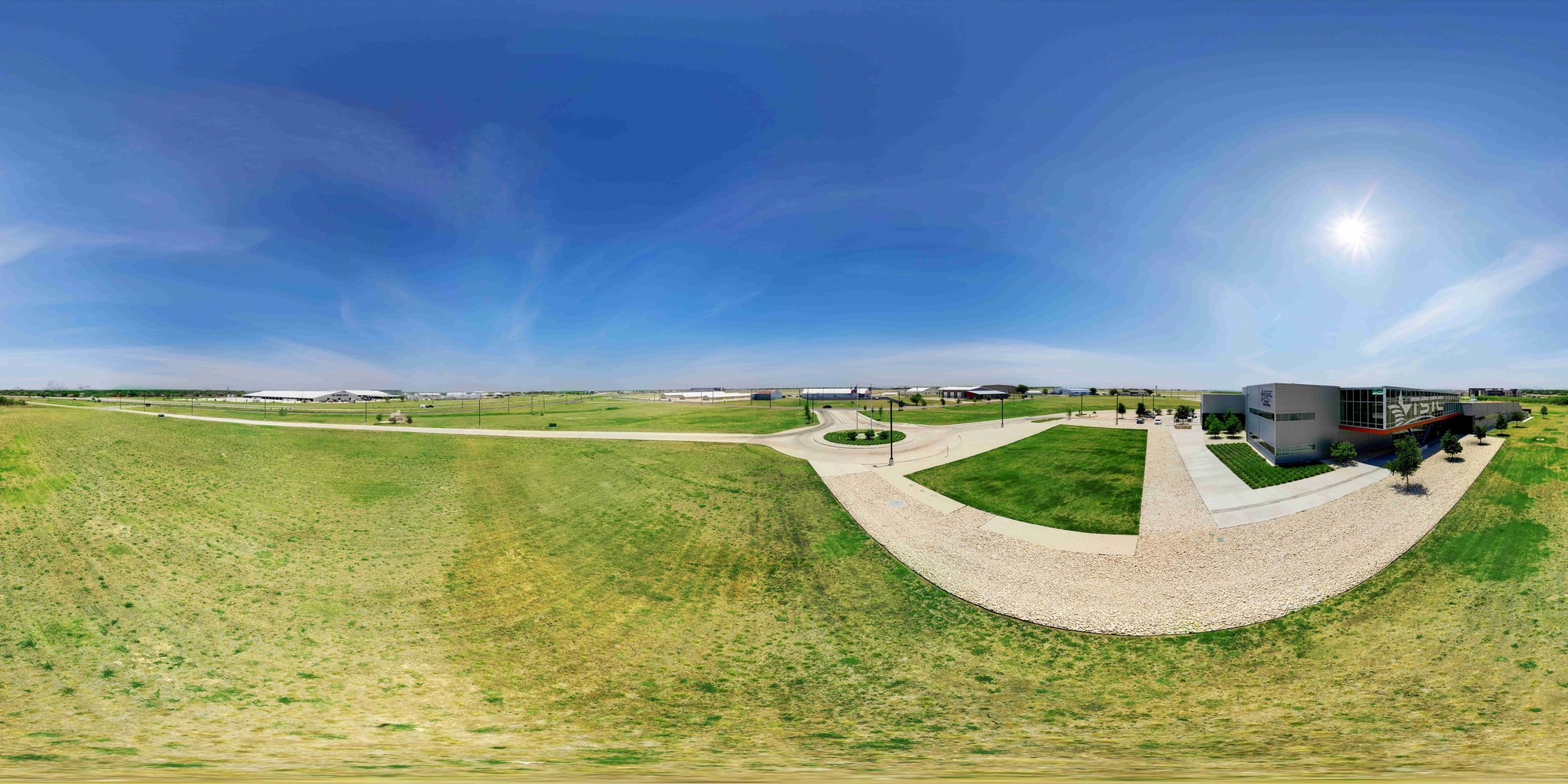 Wide panoramic view of a Texas State Technical College campus with open grassy fields, a circular drive and pathways, and a modern building on the right under a bright blue sky with the sun shining overhead.