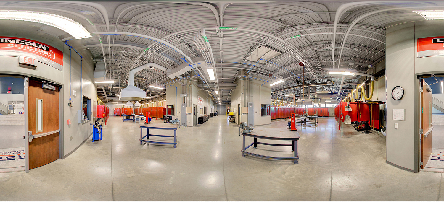 Panoramic view of an industrial training lab with welding stations enclosed by red safety curtains, metal worktables, and ventilation hoods, set within a large space with exposed piping and bright overhead lighting.