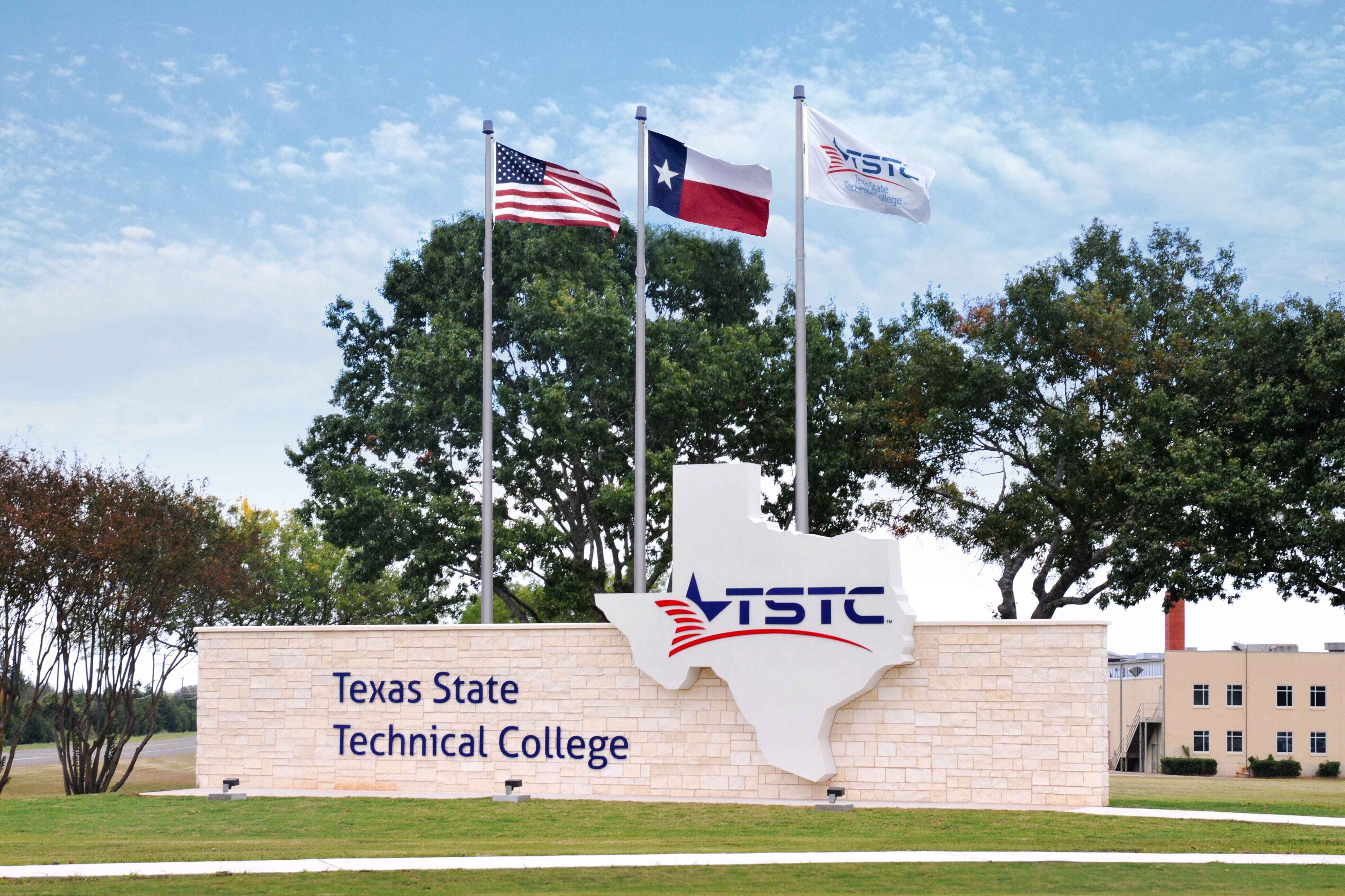 Entrance sign for Texas State Technical College featuring a large Texas-shaped emblem with the TSTC logo, flanked by three flagpoles flying the U.S., Texas, and TSTC flags, set against trees and a partly cloudy sky.