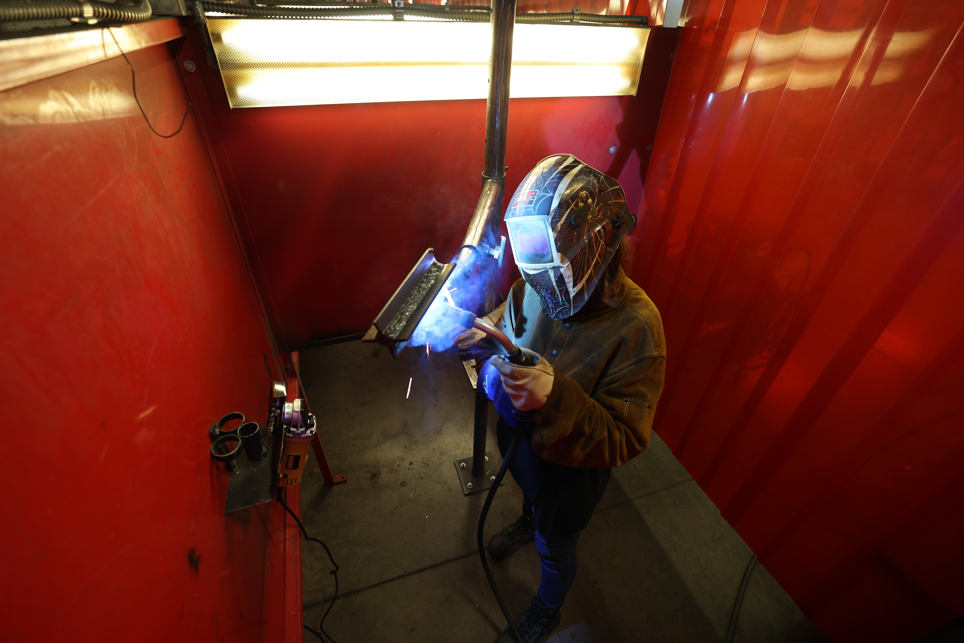 A student wearing a welding helmet and gloves performs a weld inside a red welding booth, producing bright sparks and blue arc light while holding a torch against a metal piece.