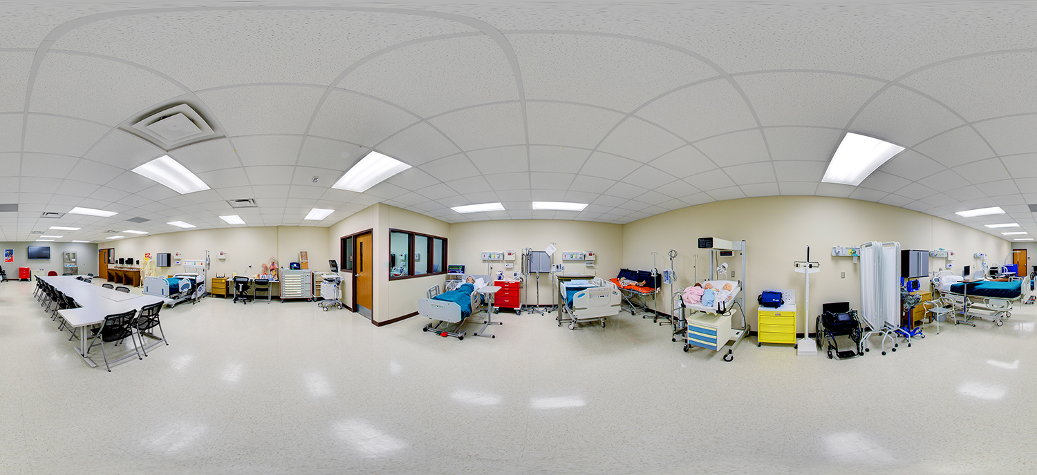 Panoramic view of a nursing skills lab with hospital beds, medical mannequins, monitoring equipment, and a classroom area with tables and chairs for instruction.