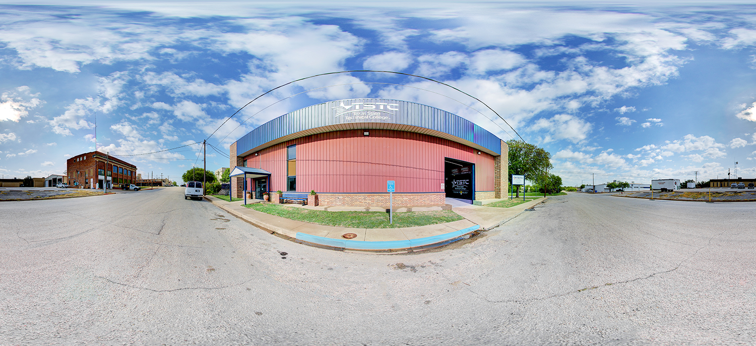 Panoramic view of a Texas State Technical College building with a curved metal roof and red exterior, situated on a street corner with surrounding roads, small-town buildings, and a partly cloudy sky.