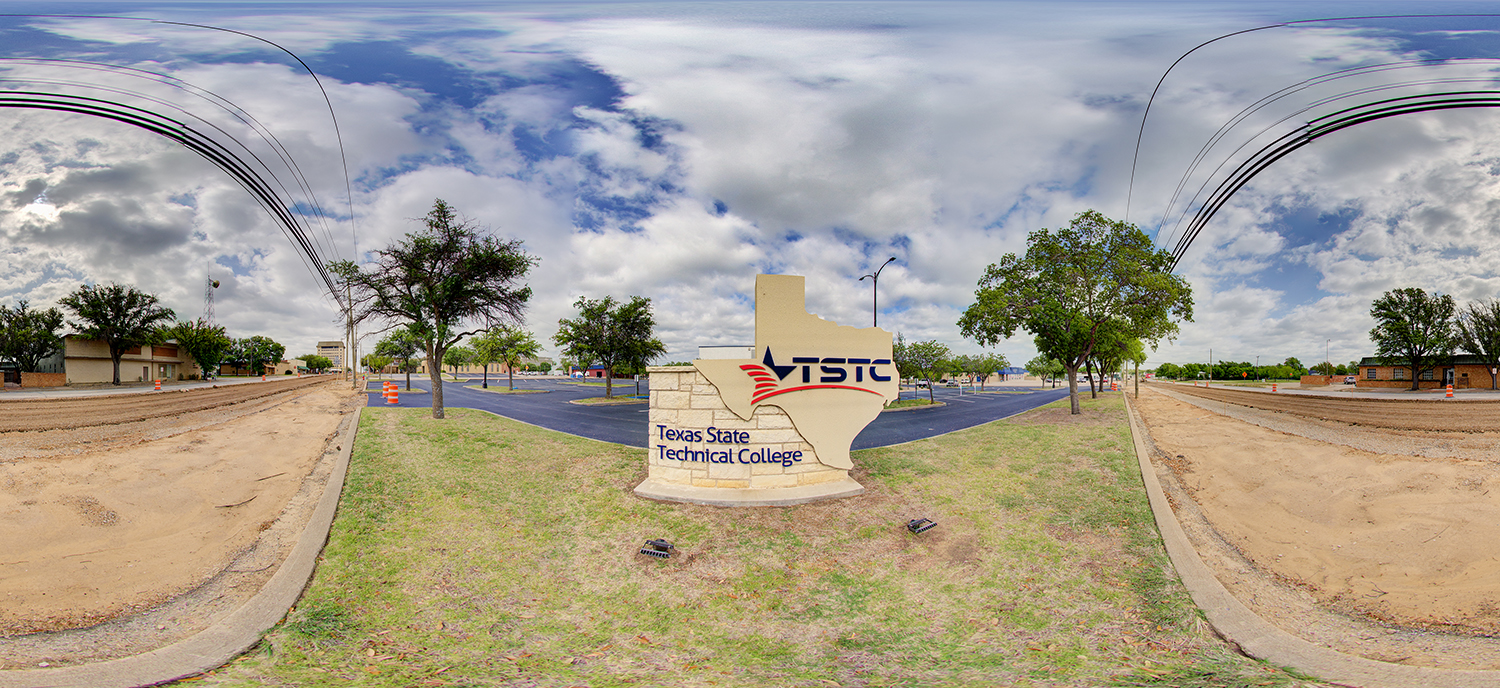 Panoramic view of a TSTC Breckenridge campus entrance sign shaped like the state of Texas, set on a grassy median with surrounding roads, trees, and a partly cloudy sky overhead.