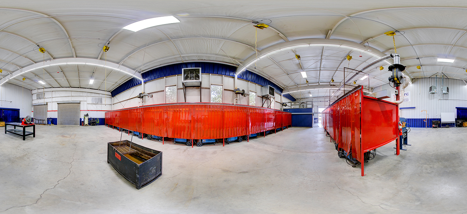 Panoramic view of a spacious welding workshop with red safety curtains enclosing work bays, overhead ventilation systems, suspended equipment, and open floor space for industrial training.