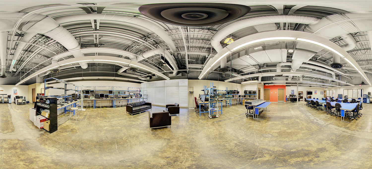 Panoramic view of a large technology lab with exposed ceiling ducts, rows of computer workstations and equipment benches, lounge seating in the center, and collaborative tables arranged along the right side.