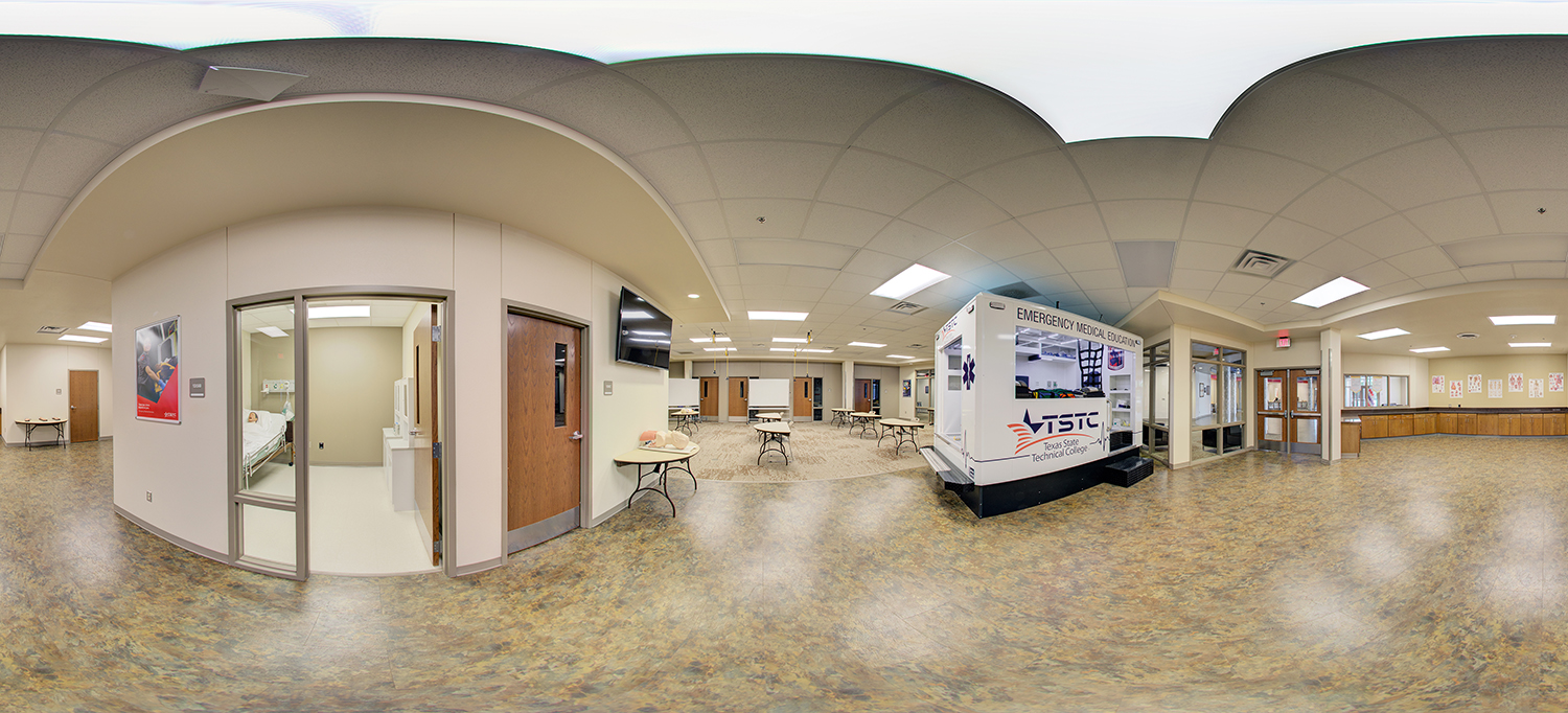 Panoramic view of a medical training classroom with hospital simulation rooms, practice mannequins on tables, and a Texas State Technical College emergency medical education training unit, surrounded by desks and instructional space.