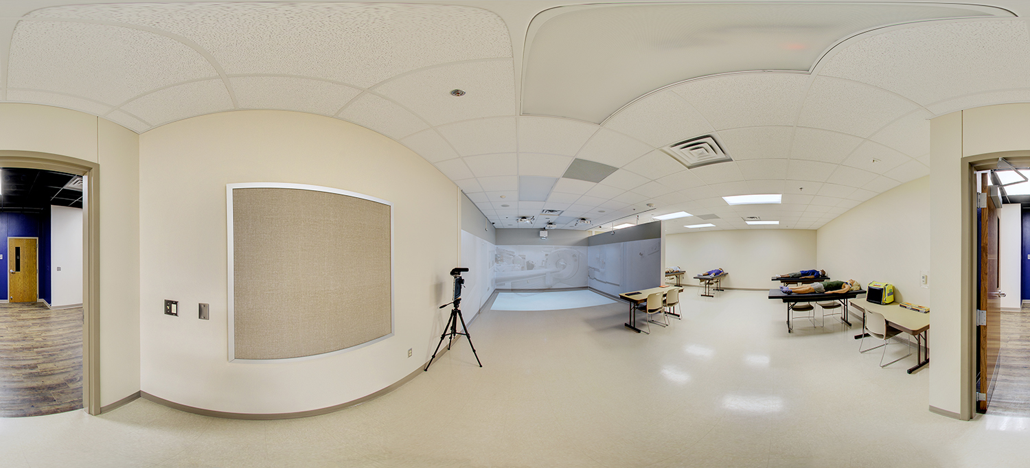 Panoramic view of a clinical training lab with medical mannequins on tables, a projection-based simulation area at the front, and classroom seating arranged around the open practice space.