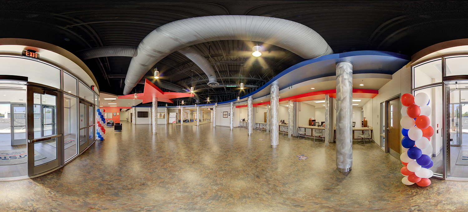 Panoramic view of a spacious campus lobby with polished floors, curved red and blue ceiling accents, rows of columns, and glass entrance doors, decorated with red, white, and blue balloon displays.