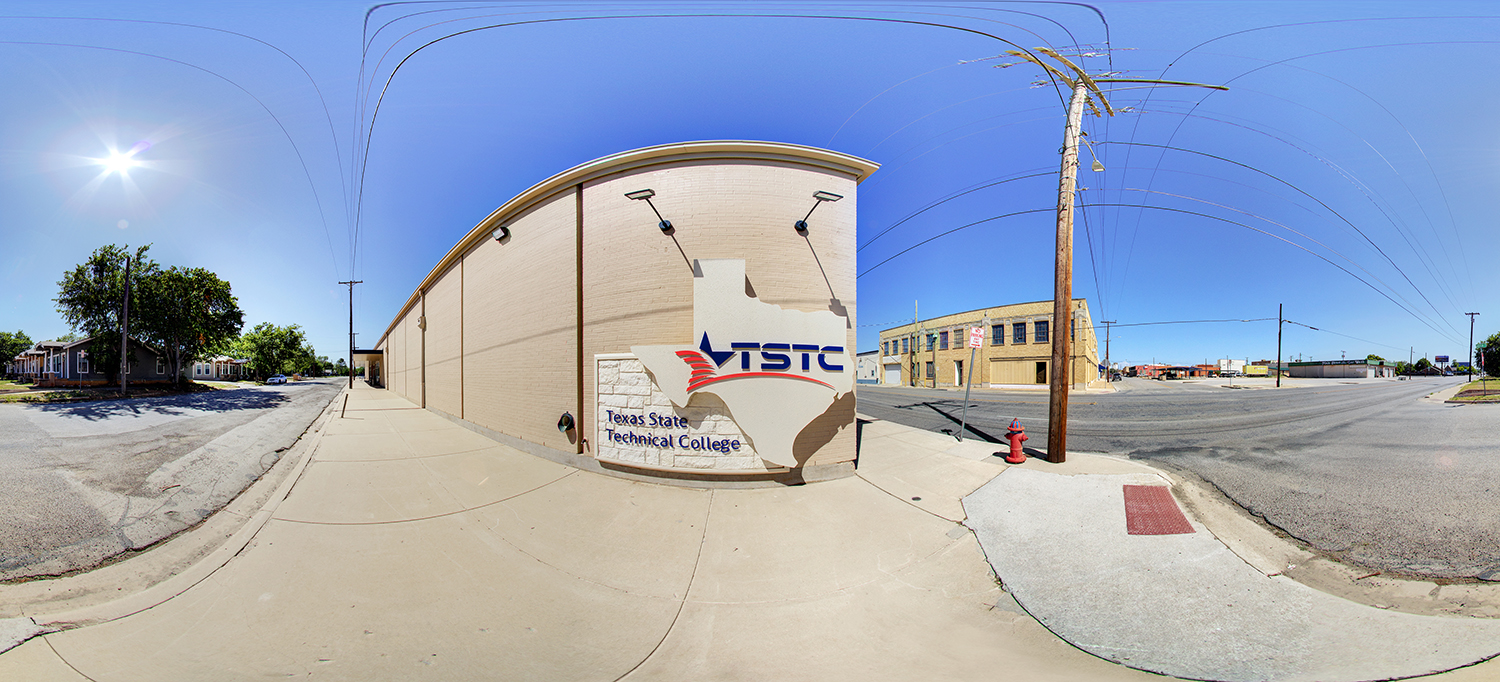 Panoramic street view of a TSTC Brownwood building with a Texas-shaped TSTC sign mounted on the exterior wall, surrounded by sidewalks, utility poles, and nearby buildings under a bright, clear sky.