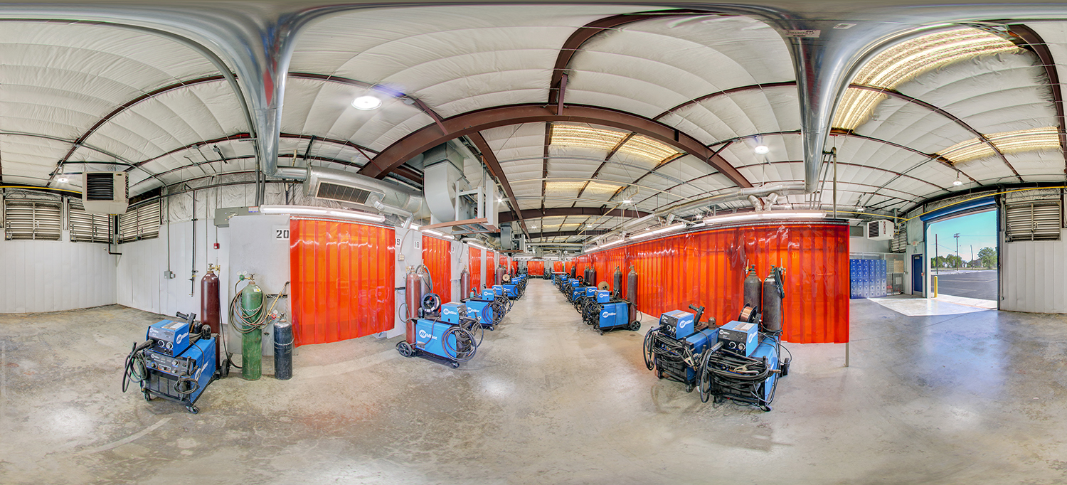 Panoramic view of an industrial welding lab with rows of blue welding machines, gas cylinders, and red translucent safety curtains dividing individual workstations inside a large, high-ceilinged shop.