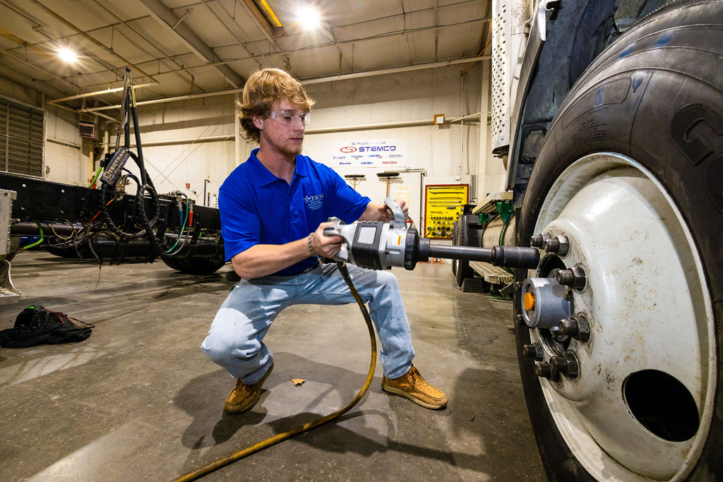 A student in safety glasses and a blue TSTC polo crouches while using a pneumatic impact wrench to loosen lug nuts on a large truck wheel inside a mechanic training workshop.