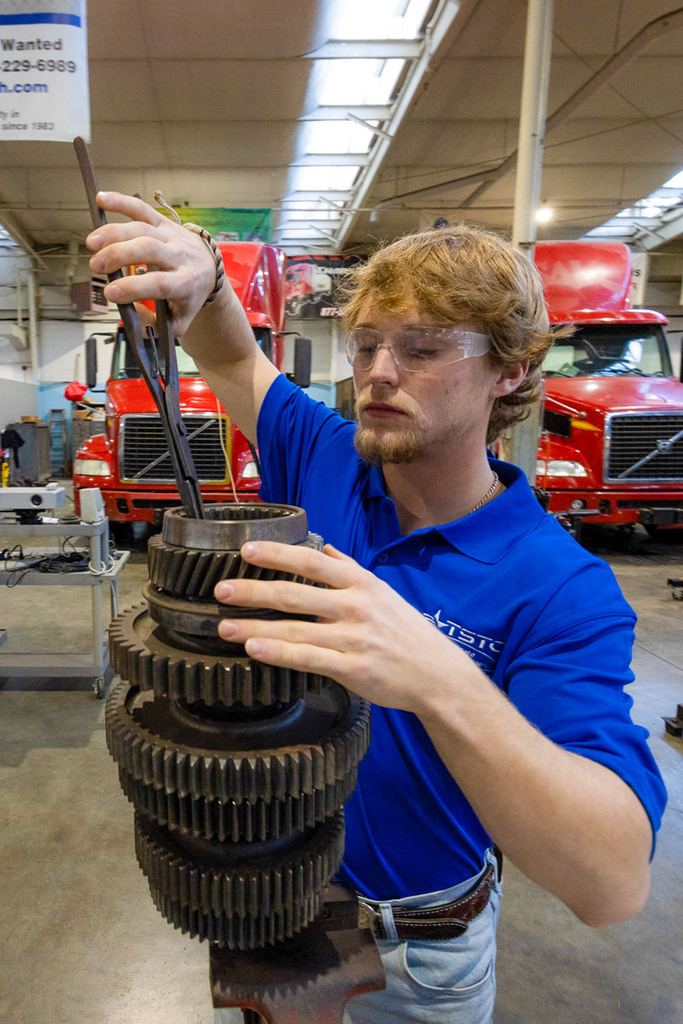 A student wearing safety glasses and a blue TSTC polo carefully lifts and inspects stacked metal gears using pliers in an automotive lab with trucks in the background.