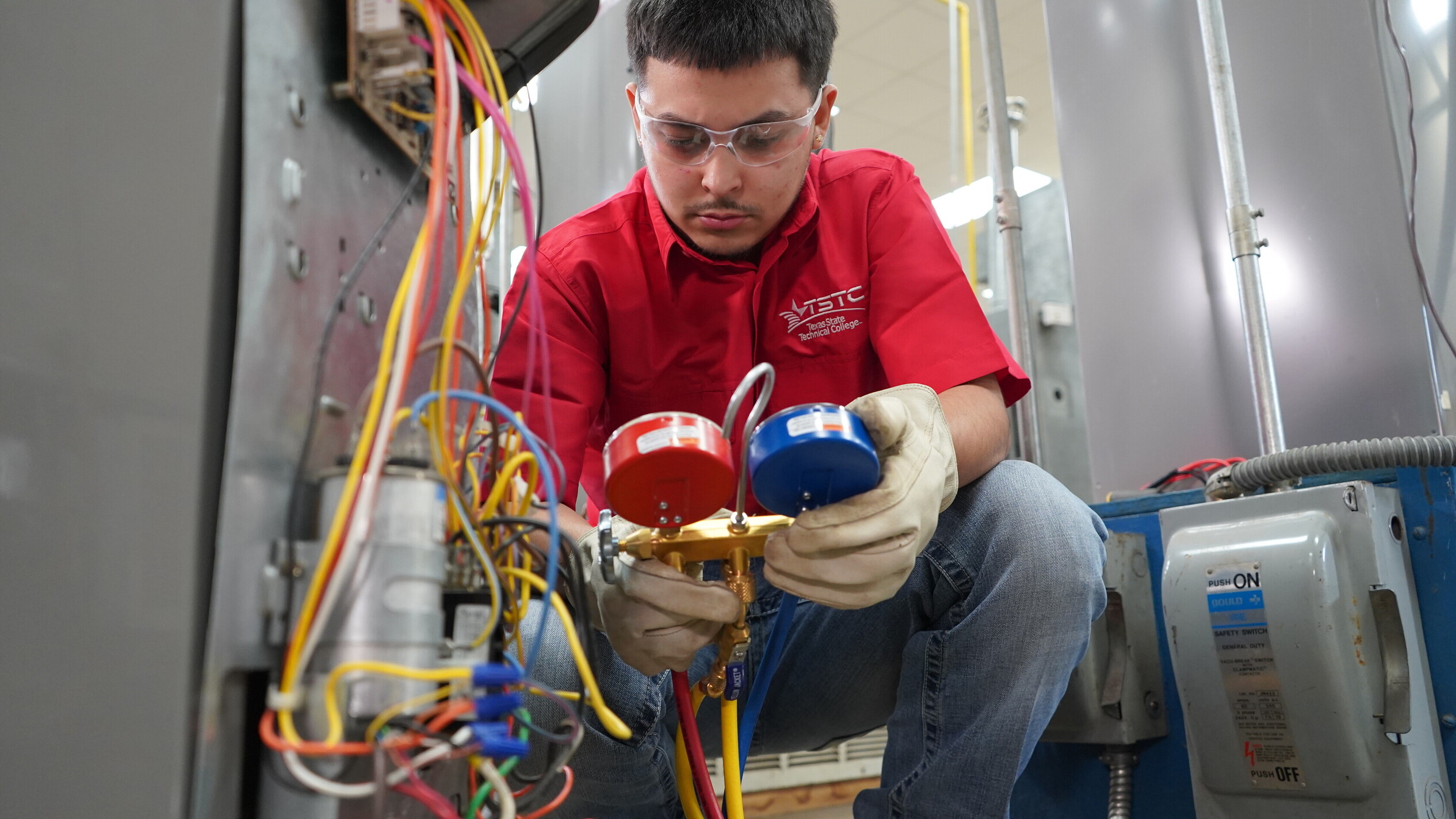 A student in safety glasses and a red TSTC shirt crouches while using manifold gauges to measure pressure in an HVAC system, surrounded by exposed wiring and equipment.