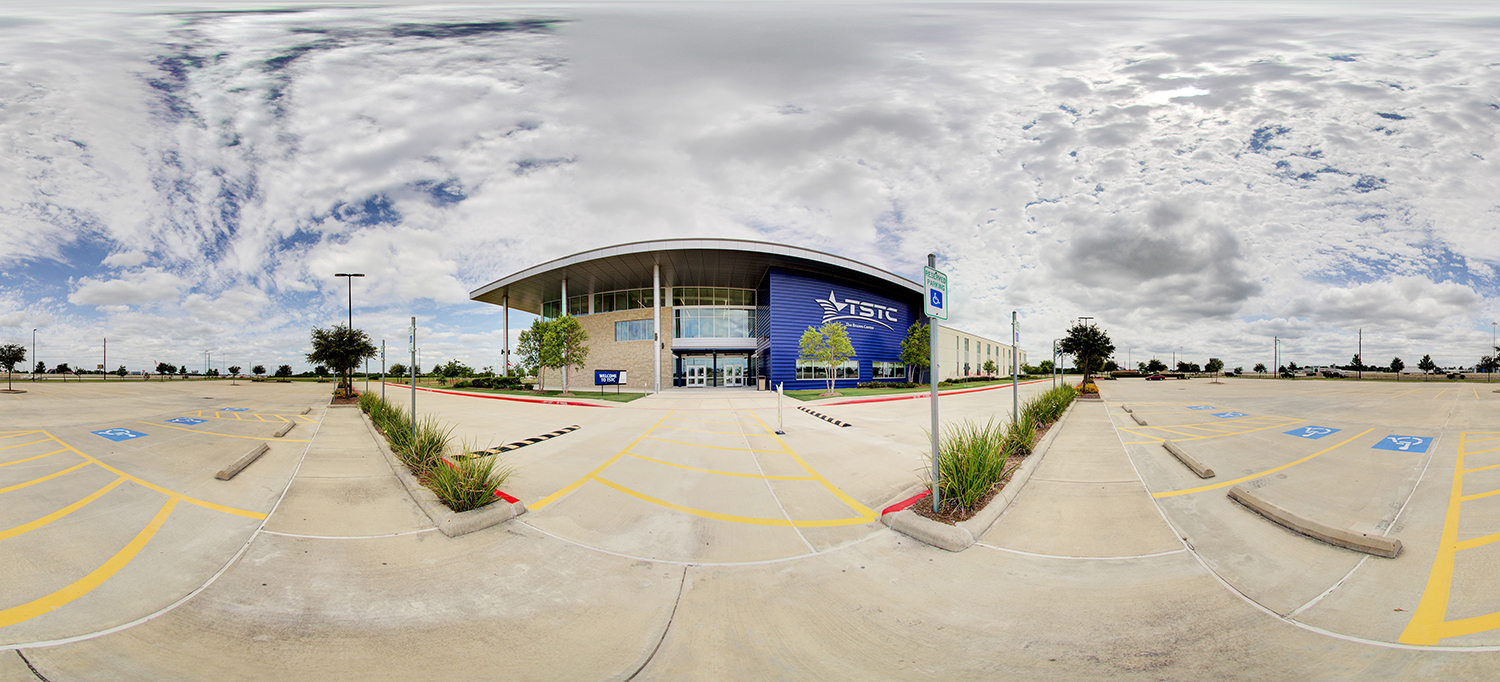 Panoramic view of a Texas State Technical College building with a curved roof and blue facade, surrounded by a mostly empty parking lot with accessible parking spaces, small landscaped areas, and a cloudy sky overhead.