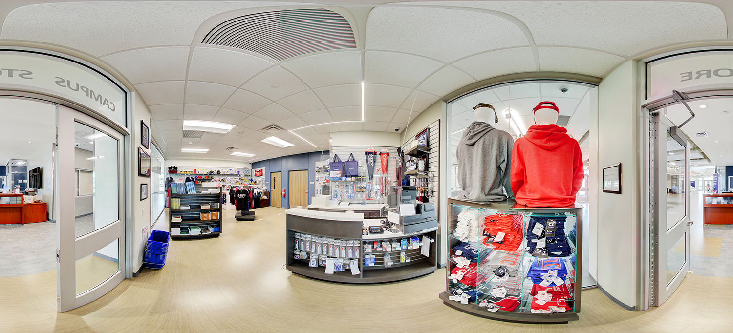 Panoramic view of a campus store interior with display racks of clothing and merchandise, a central checkout counter, and mannequins wearing hoodies, all arranged in a bright, retail-style space.