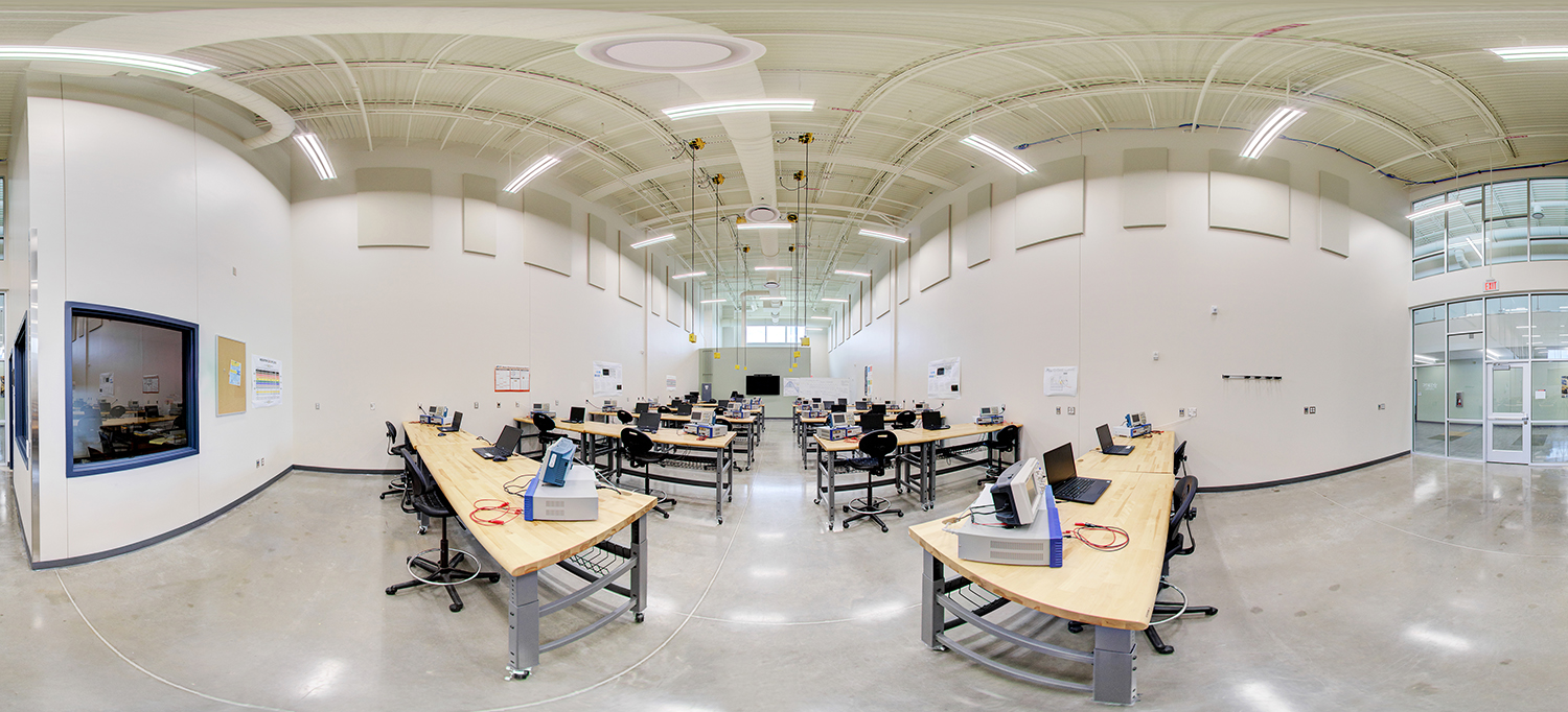 Panoramic view of a spacious electronics or engineering lab with high ceilings, rows of wooden workbenches equipped with testing devices and laptops, rolling stools, and instructional materials on the walls.