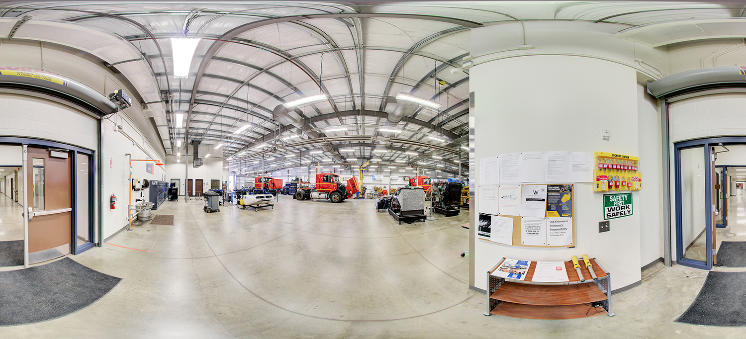 Panoramic view of a large automotive or heavy equipment training workshop with high ceilings, where multiple red trucks and machinery are arranged across an open floor, surrounded by tools, workstations, and safety signage on nearby walls.