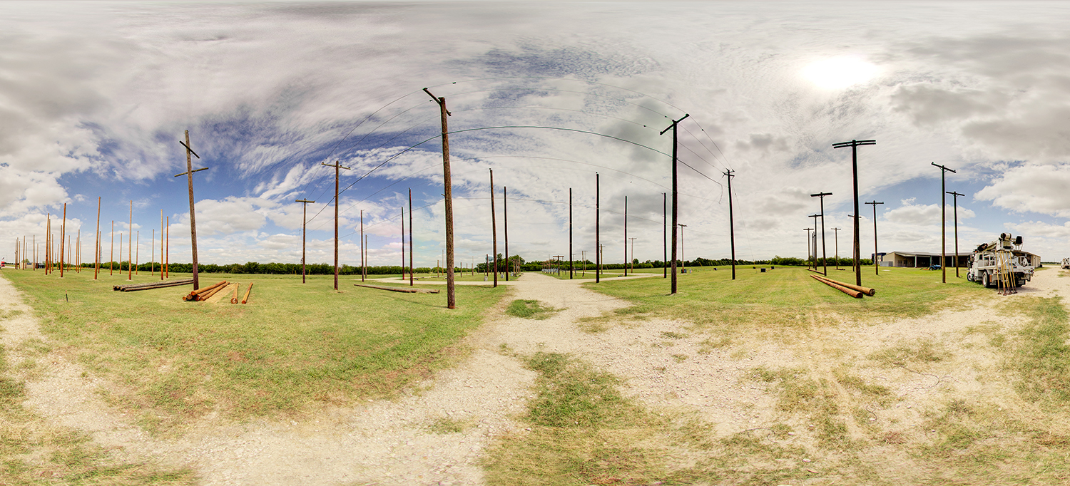 Panoramic view of an outdoor electrical lineworker training yard with numerous utility poles—some connected by overhead wires—set across a grassy field, with dirt paths, stacked poles on the ground, and service trucks and a small building nearby under a partly cloudy sky.