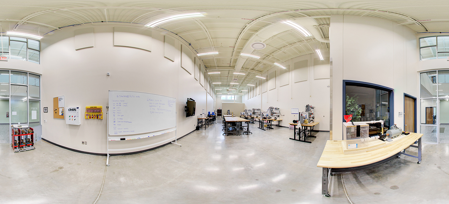 Panoramic view of a technical training lab with high ceilings and bright lighting, featuring workbenches with machinery and equipment, a large whiteboard with notes, and instructional materials arranged along the walls.