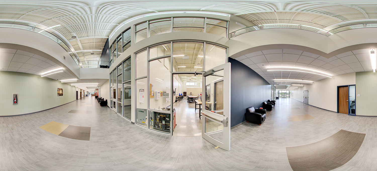 Panoramic view of a modern academic hallway with light gray flooring and high ceilings, featuring a glass-walled lab classroom with an open door at center, rows of black lounge chairs along the walls, and bright overhead lighting throughout
