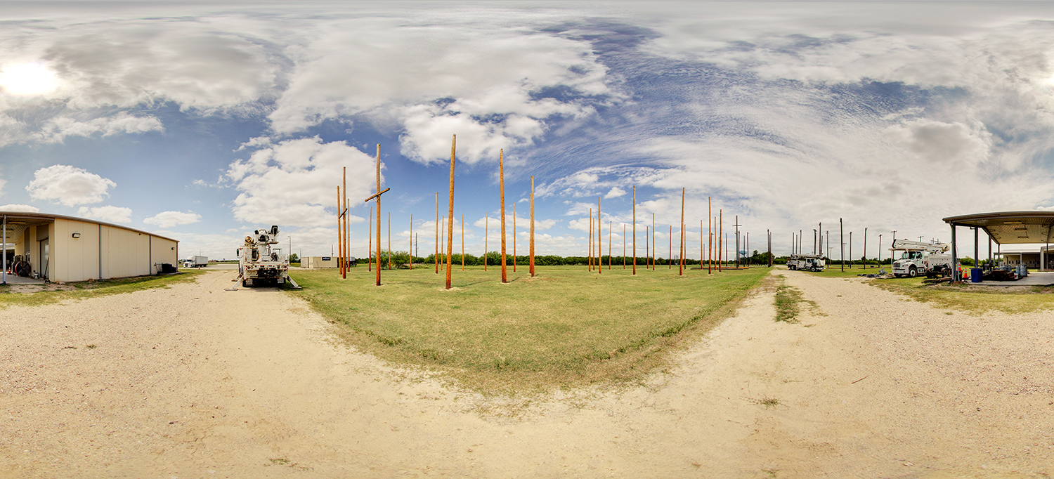 Panoramic view of an outdoor utility line training field with numerous tall wooden poles arranged in rows, surrounded by grassy terrain and dirt paths, with service trucks and equipment parked near adjacent buildings under a partly cloudy sky.
