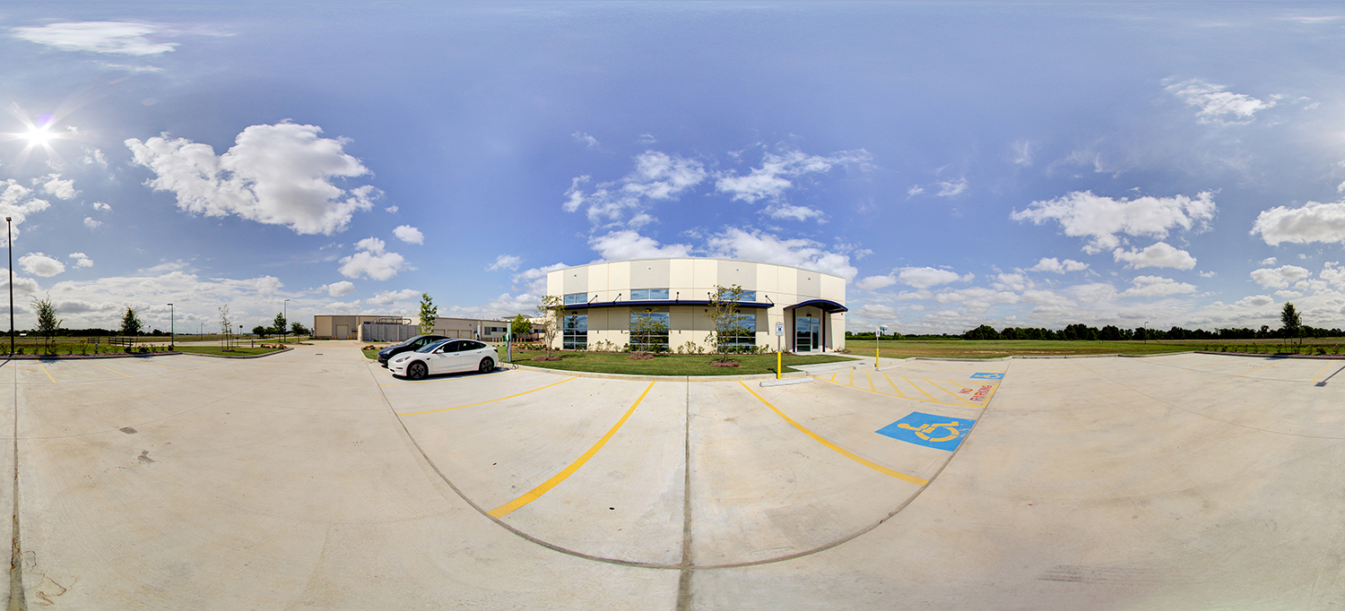 Panoramic view of a modern campus building with a curved facade and large windows, surrounded by a spacious parking lot with marked accessible spaces, a few parked cars, and landscaped areas under a bright blue sky with scattered clouds.
