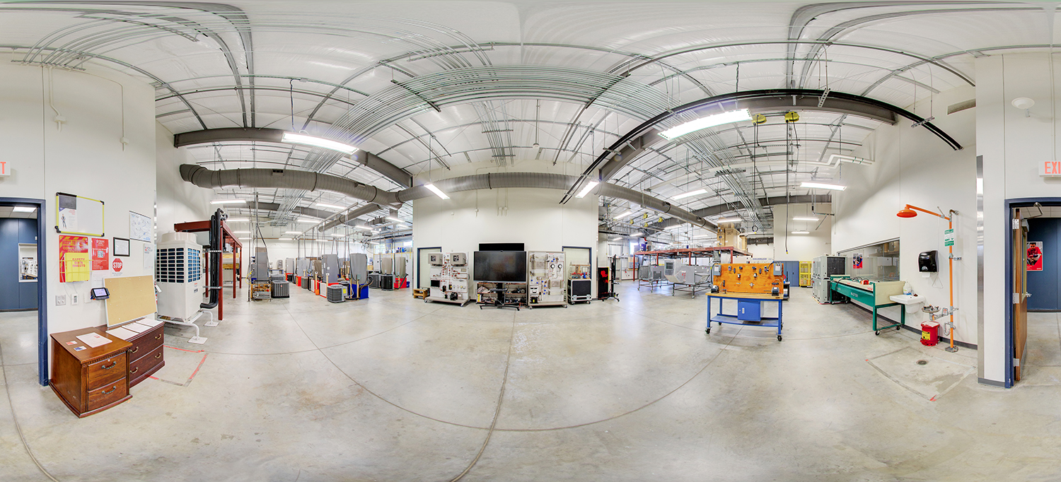 Panoramic view of an industrial training lab with high ceilings and exposed ductwork, featuring HVAC units, control panels, and technical equipment arranged around the room, with workbenches, safety stations, and instructional displays along the walls.