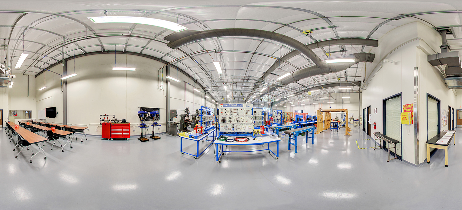 Panoramic view of an industrial training lab with high ceilings and exposed ductwork, featuring motor control system panels, conveyor setups, tool stations, and rows of classroom seating along one side under bright overhead lighting.