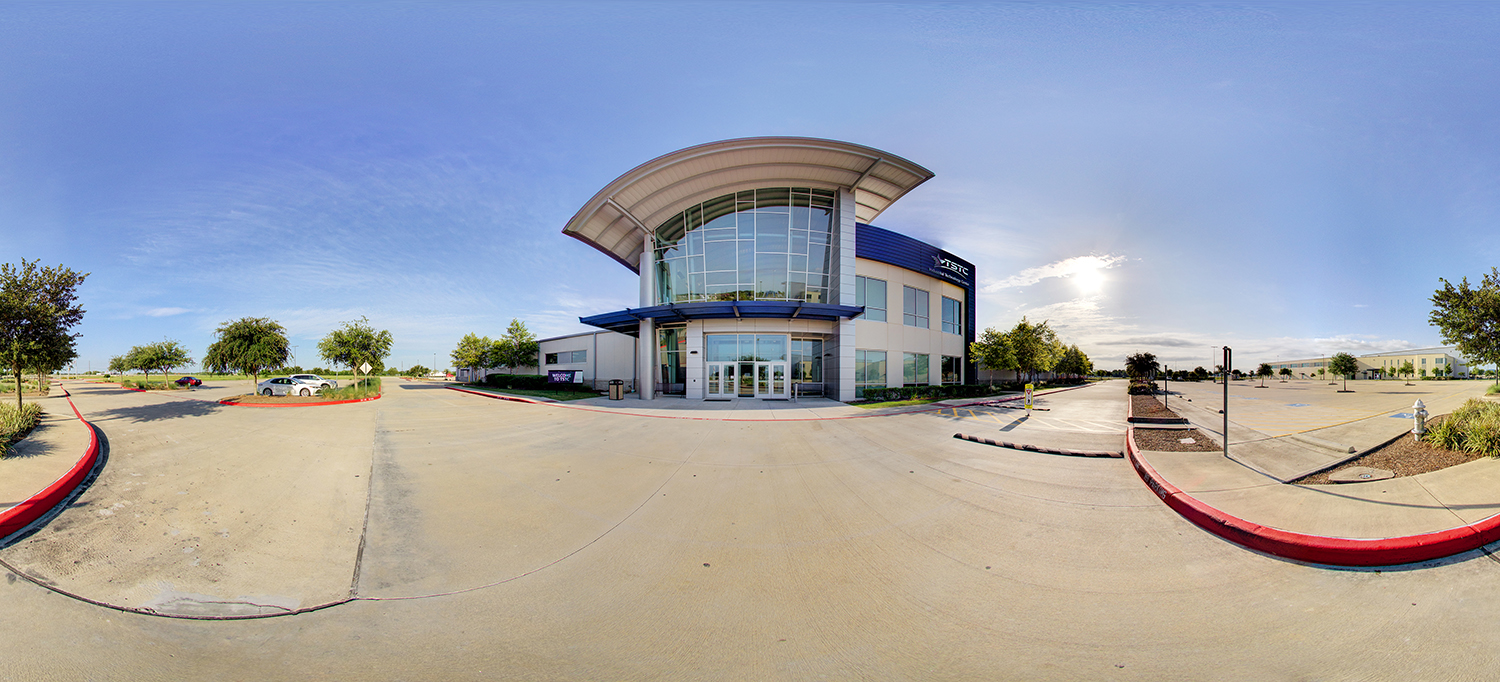 Panoramic view of a modern Texas State Technical College building with a curved roof and glass entrance, surrounded by a wide driveway, parking areas, landscaped islands, and a clear blue sky with sunlight.