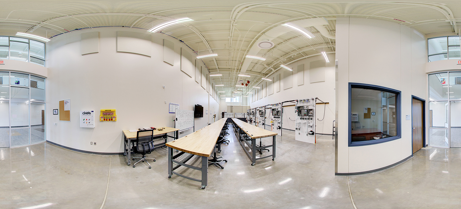 Panoramic view of a technical training classroom with high ceilings, long wooden workbenches arranged in rows, rolling stools, wall-mounted electrical training panels, and a whiteboard with notes.