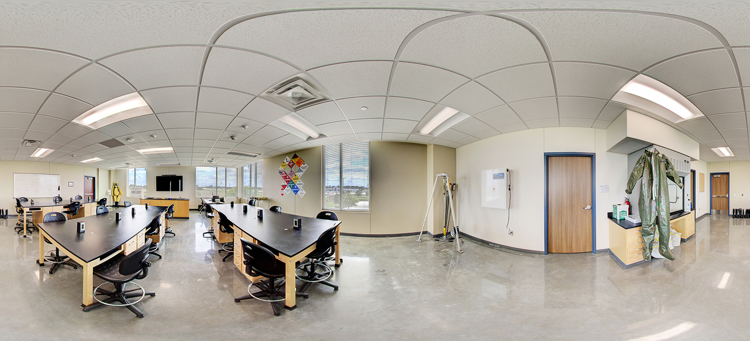 Panoramic view of a science or safety training lab with long black worktables and stools, large windows providing natural light, wall-mounted equipment, and protective suits hanging near storage cabinets.