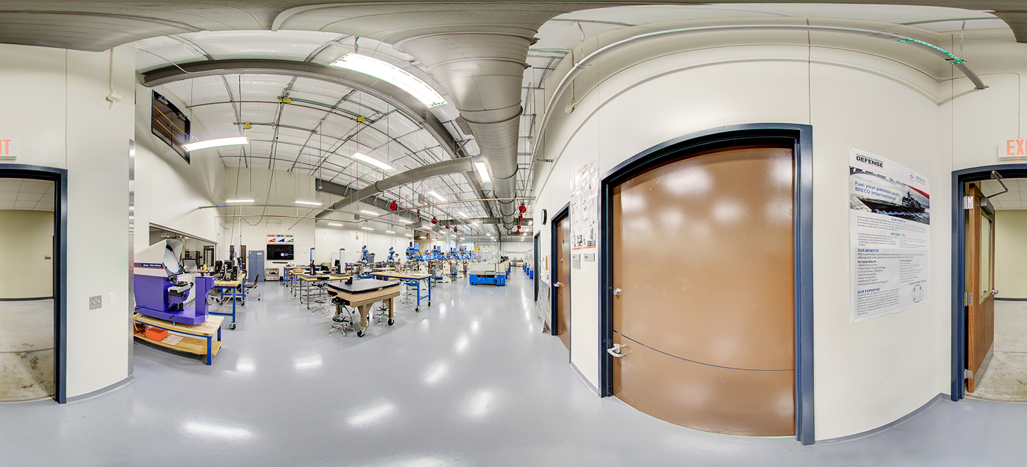 Panoramic view of a manufacturing or machining training lab with polished floors and high ceilings, showing rows of precision equipment, worktables, and measurement stations, with instructional posters and doors lining the perimeter walls.